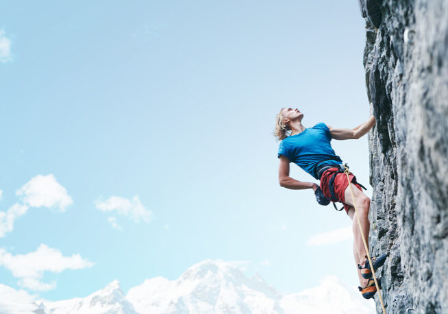 man rock climber with long hair. side view of young man rock climber in bright red shorts climbing the challenging route on the cliff on the blue sky background. rock climber climbs on a rocky wall.